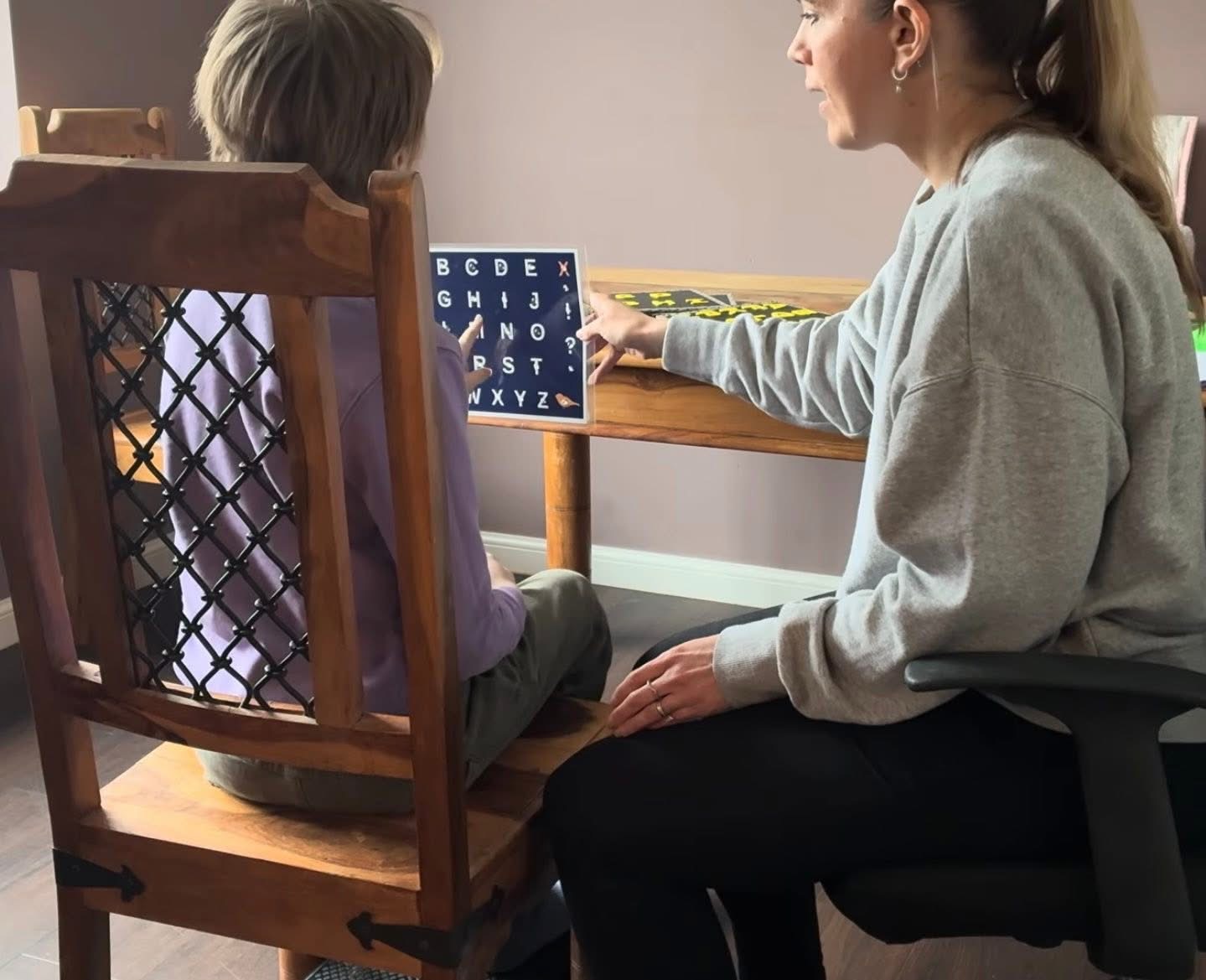 Lydia sitting at a table with a speller who is pointing to a letterboard during an S2C session.